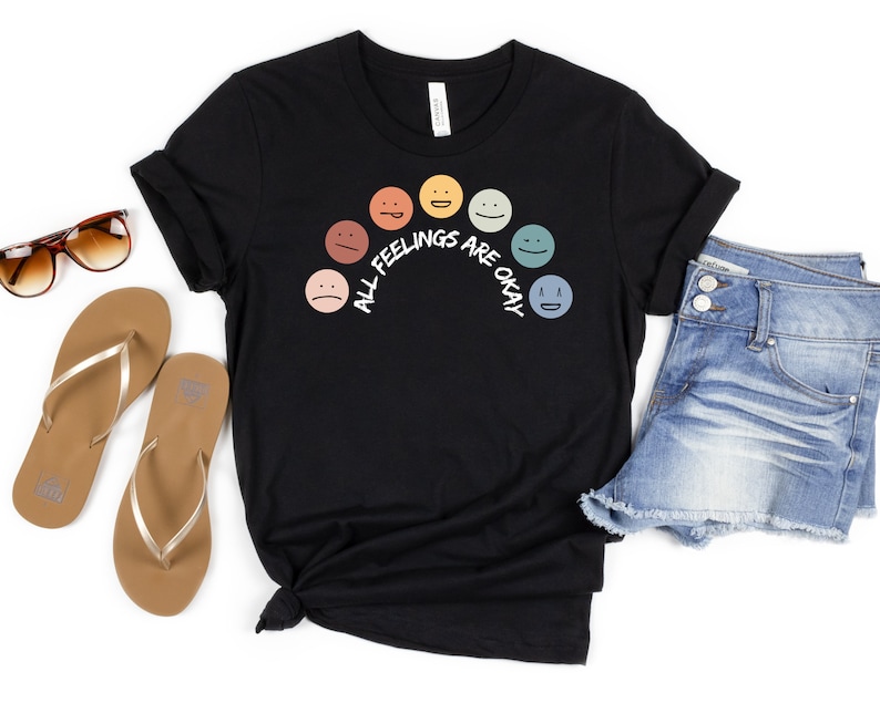 Op de afbeelding: Zwart T-shirt met een regenboog van kleurrijke gezichten en de tekst "Alle gevoelens zijn ok&eacute;".