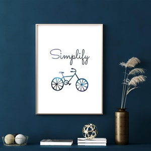 May include: A white framed print with the word "Simplify" in a blue gradient font and a black silhouette of a bicycle.