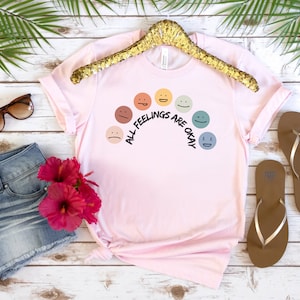 Op de afbeelding: Roze T-shirt met een regenboog van kleurrijke gezichten en de tekst "Alle gevoelens zijn ok&eacute;".