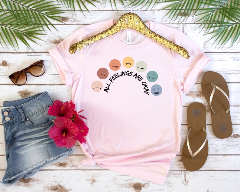 Op de afbeelding: Roze T-shirt met een regenboog van kleurrijke gezichten en de tekst "Alle gevoelens zijn ok&eacute;".