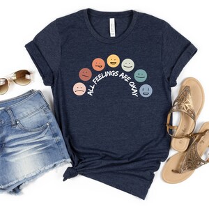 Op de afbeelding: Een marineblauw t-shirt met een regenboog van kleurrijke gezichten en de tekst "Alle gevoelens zijn ok&eacute;".