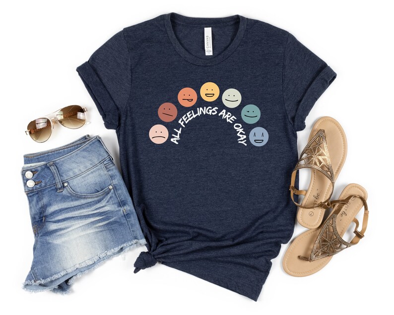 Op de afbeelding: Een marineblauw t-shirt met een regenboog van kleurrijke gezichten en de tekst "Alle gevoelens zijn ok&eacute;".