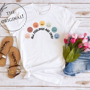 Op de afbeelding: Wit T-shirt met een regenboog van kleurrijke gezichten en de tekst "Alle gevoelens zijn ok&eacute;".