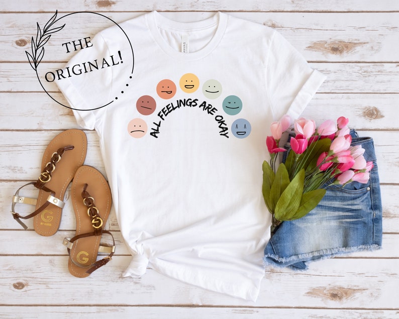 Op de afbeelding: Wit T-shirt met een regenboog van kleurrijke gezichten en de tekst "Alle gevoelens zijn ok&eacute;".