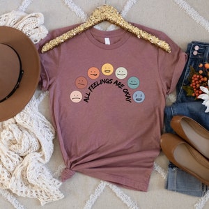 Op de afbeelding: Een mauve t-shirt met een regenboog van kleurrijke smiley-gezichten en de tekst "All Feelings Are Okay".