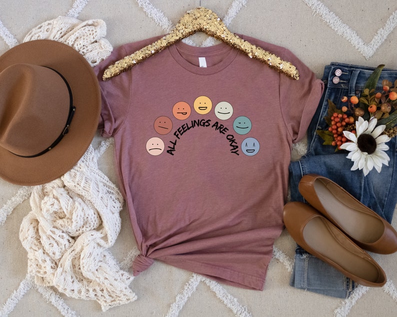 Op de afbeelding: Een mauve t-shirt met een regenboog van kleurrijke smiley-gezichten en de tekst "All Feelings Are Okay".