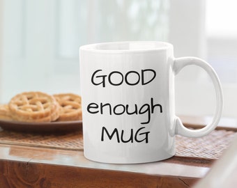Op de afbeelding: Een witte keramische mok met zwarte tekst die "GOOD enough MUG" luidt. De mok staat op een houten tafel.