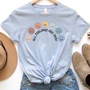 Op de afbeelding: Een lichtblauw t-shirt met een regenboog van verschillende gekleurde gezichten en de tekst "All Feelings Are Okay".