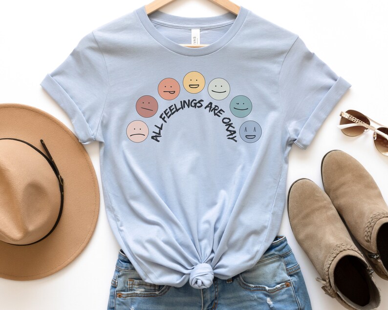 Op de afbeelding: Een lichtblauw t-shirt met een regenboog van verschillende gekleurde gezichten en de tekst "All Feelings Are Okay".