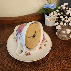 May include: A white teacup and saucer with a floral rose pattern, repurposed as a clock. The clock face is made of wood, with black hands and hour markers. The teacup is resting on its side on the saucer.