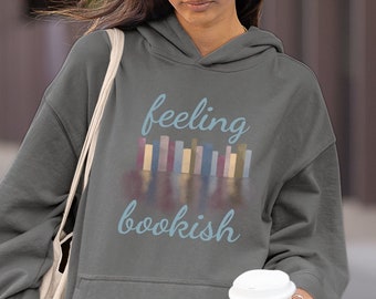 May include: A charcoal gray hooded sweatshirt featuring a graphic of colorful book spines and the text "feeling bookish" in light blue script. The hoodie includes a front kangaroo pocket and is paired with a matching charcoal gray baseball cap.
