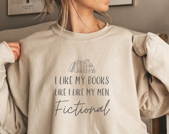 May include: A beige crewneck sweatshirt featuring a graphic of a row of books and the text "I LIKE MY BOOKS LIKE I LIKE MY MEN Fictional" printed in dark gray ink across the chest. The garment has long sleeves and a ribbed neckline.