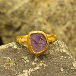 May include: A gold ring featuring a purple gemstone. The ring is displayed on a rough, textured rock.