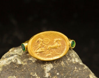May include: A gold ring with an oval-shaped bezel featuring an engraved design of a deer. The ring is accented with two green gemstones on either side of the bezel.