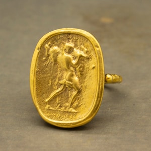 May include: A gold-toned ring with an oval-shaped face featuring a raised design of a person holding a staff. The ring band is textured.