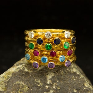 Può includere: Un set di anelli d'oro con pietre preziose colorate. Gli anelli sono disposti in cerchio, con le pietre preziose rivolte verso l'esterno. Le pietre preziose sono di vari colori, tra cui blu, verde, rosso e viola.