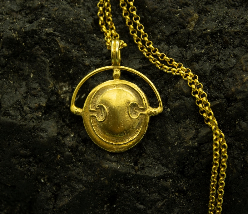 Puede incluir: Un collar de oro con un colgante redondo y adornado con un dise&ntilde;o estilizado. El colgante est&aacute; suspendido de una delicada cadena de oro.