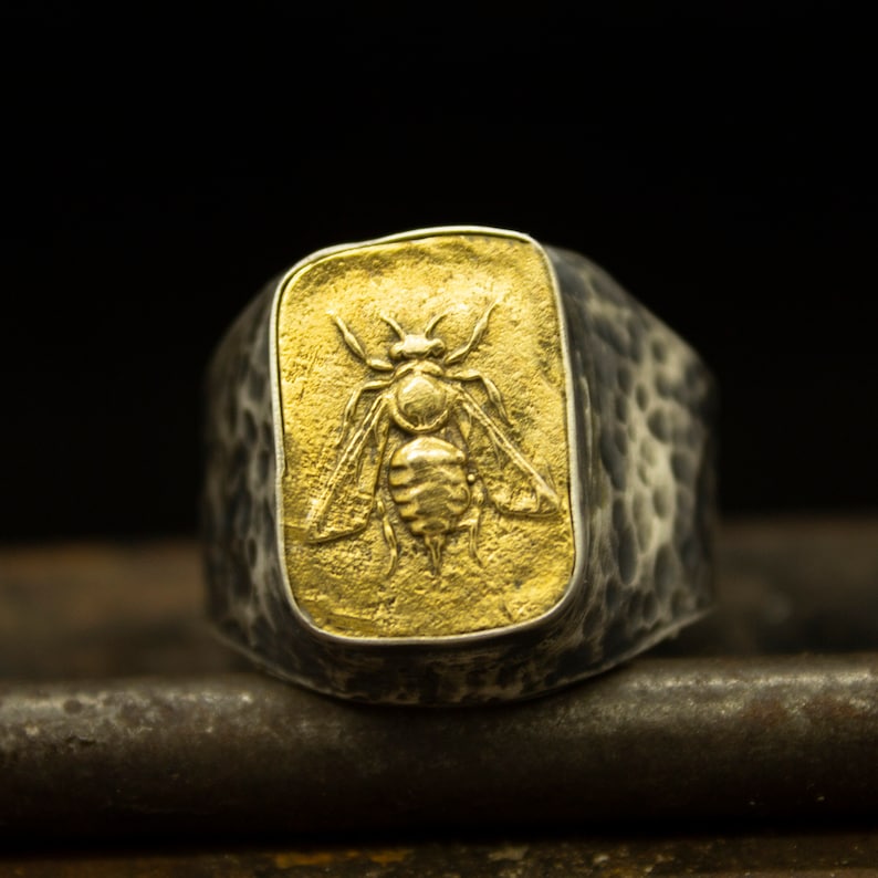 May include: A silver ring with a textured band and a rectangular gold inset featuring a detailed bee design.