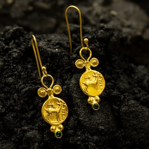 Pu&ograve; includere: Un paio di orecchini pendenti in oro con un accento di pietra preziosa verde. Ogni orecchino presenta un ciondolo rotondo con un'immagine dettagliata di un cervo.