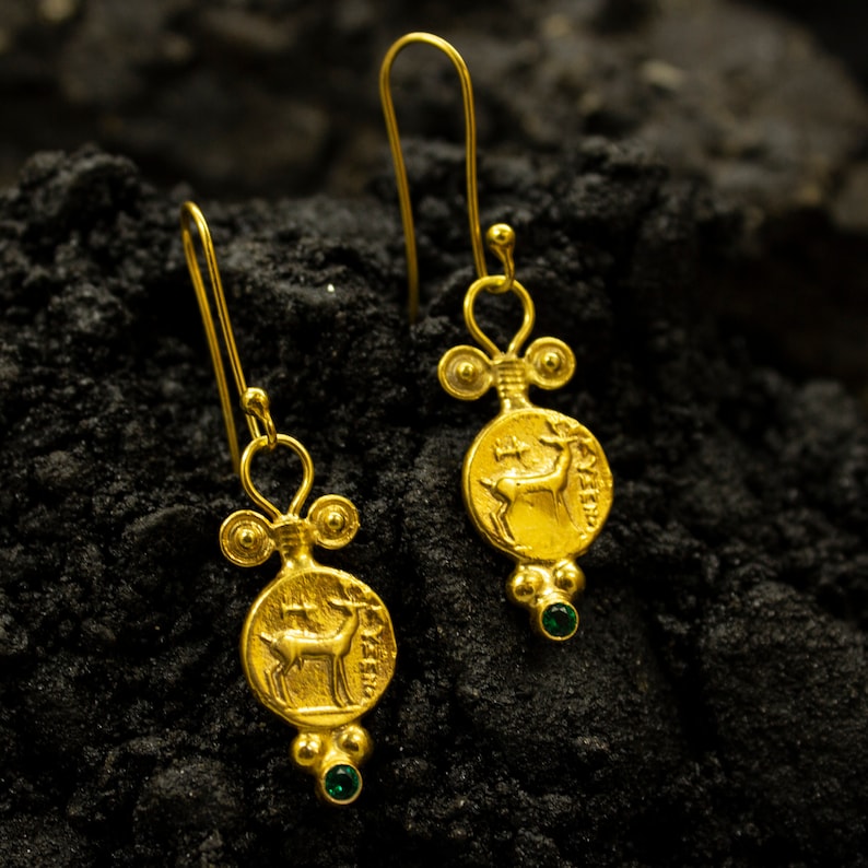 Pu&ograve; includere: Un paio di orecchini pendenti in oro con un accento di pietra preziosa verde. Ogni orecchino presenta un ciondolo rotondo con un'immagine dettagliata di un cervo.