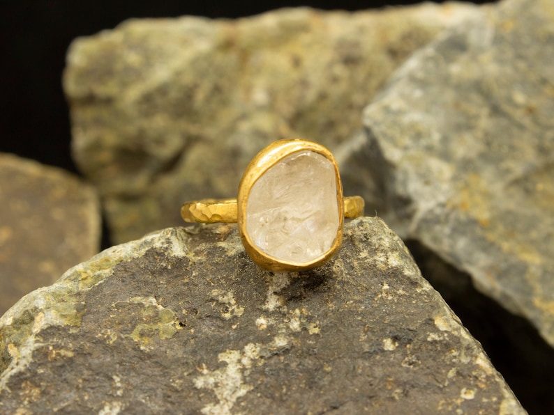 May include: A gold ring with a clear, oval-shaped gemstone. The ring is set on a rough, gray rock.