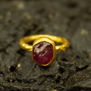Pode incluir: Um anel de ouro com uma gema facetada de cor vermelha escura engastada em uma banda texturizada.