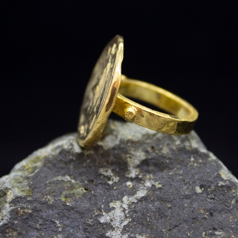 Peut inclure: Une bague en or avec un grand visage rond et textur&eacute;. La bague est pos&eacute;e sur une roche gris fonc&eacute;.