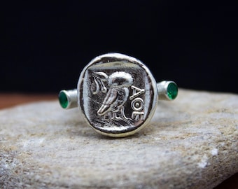 May include: A silver ring with a round, raised design featuring a bird and the inscription "AOE". The ring has two green gemstones on the band.