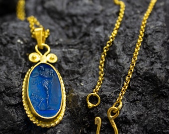 May include: A gold-colored necklace with a blue oval pendant. The pendant features a detailed figure and is framed by a twisted gold border. The necklace chain and clasp are also gold, set against a dark, textured background.