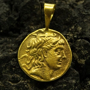 May include: A gold pendant featuring a detailed profile of a person with flowing hair. The pendant is on a dark, textured surface.