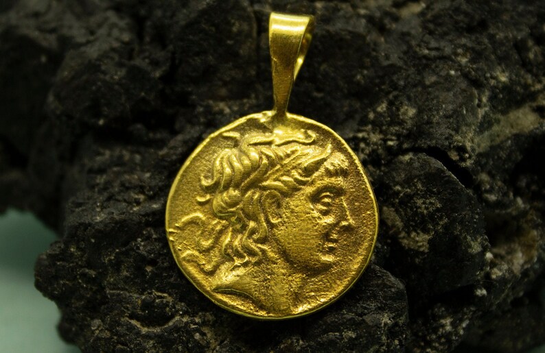 May include: A gold pendant featuring a detailed profile of a person with flowing hair. The pendant is on a dark, textured surface.