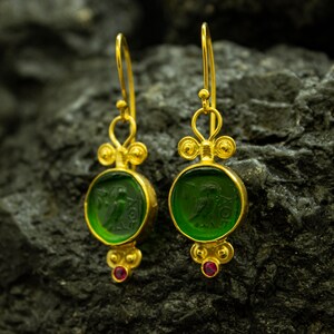 May include: Gold earrings with green glass insets featuring a carved design. The earrings have a small red gemstone at the bottom.
