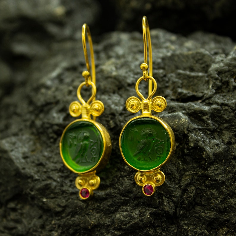 May include: Gold earrings with green glass insets featuring a carved design. The earrings have a small red gemstone at the bottom.