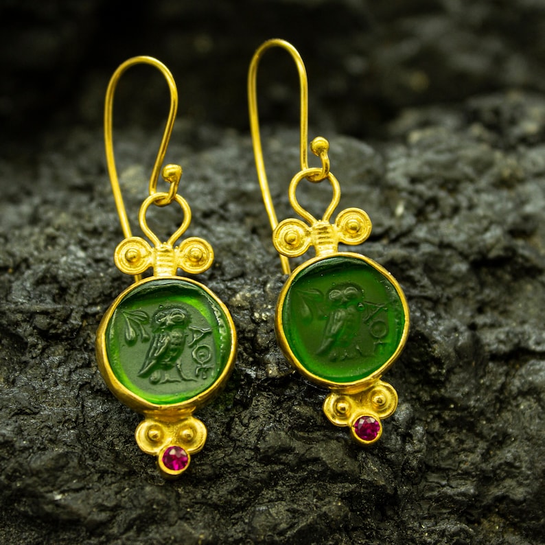 May include: Gold earrings with green glass insets featuring an owl design. The earrings have a dangling design with small red gemstones at the bottom.