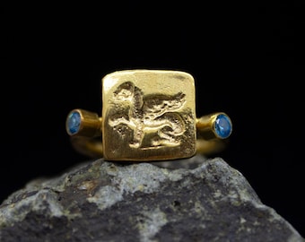 May include: Gold ring with a square-shaped bezel featuring a detailed engraving of a winged creature. The ring is accented with two small blue gemstones on either side of the bezel.