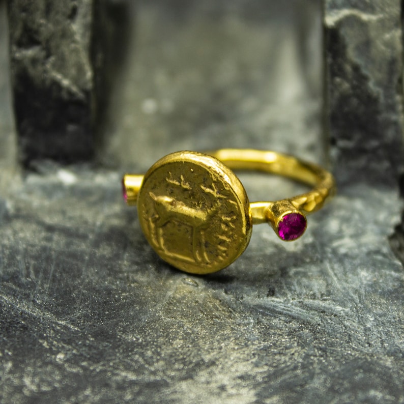 Puede incluir: Anillo de oro con un centro redondo en forma de moneda que presenta una imagen grabada de un ciervo. El anillo tiene dos peque&ntilde;as piedras preciosas rosadas a cada lado de la moneda.