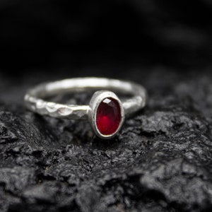 Può includere: Un anello d'argento con una fascia martellata e una vivace pietra preziosa rossa a taglio ovale. L'anello è presentato su uno sfondo scuro e strutturato, evidenziando il contrasto tra la pietra brillante e il metallo.