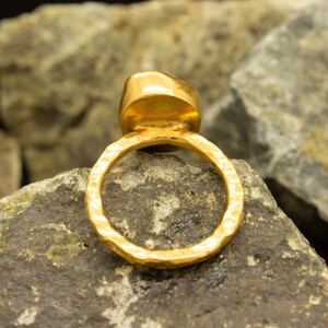 May include: A gold ring with a hammered band and a large, round, gold gemstone. The ring is sitting on a rock.