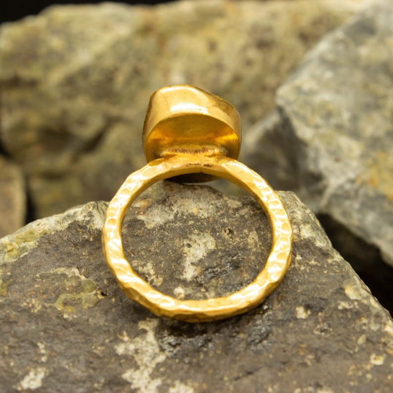 May include: A gold ring with a hammered band and a large, round, gold gemstone. The ring is sitting on a rock.