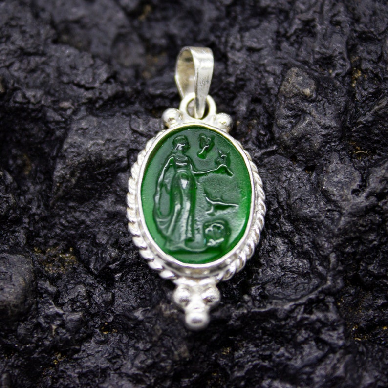 Puede incluir: Un colgante de plata con un camafeo verde que representa a una mujer sosteniendo un p&aacute;jaro. El camafeo est&aacute; engastado en un marco ovalado de plata con un dise&ntilde;o de cuerda.