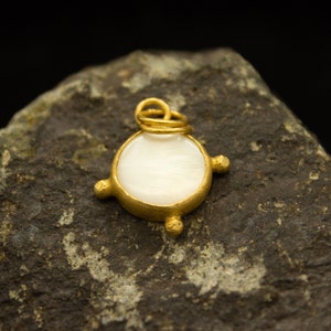 May include: A gold pendant with a white mother-of-pearl inlay. The pendant is round and has a simple design with four small gold accents around the edge.
