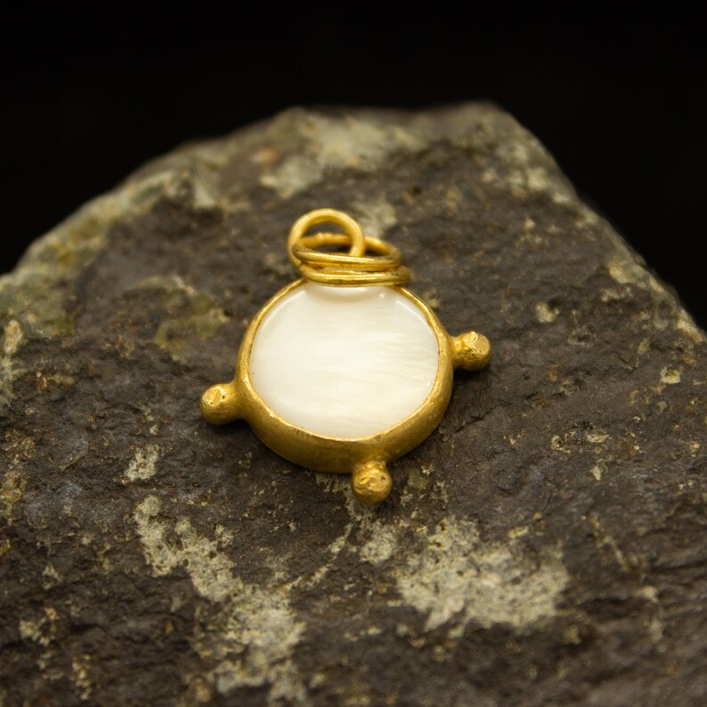 May include: A gold pendant with a white mother-of-pearl inlay. The pendant is round and has a simple design with four small gold accents around the edge.