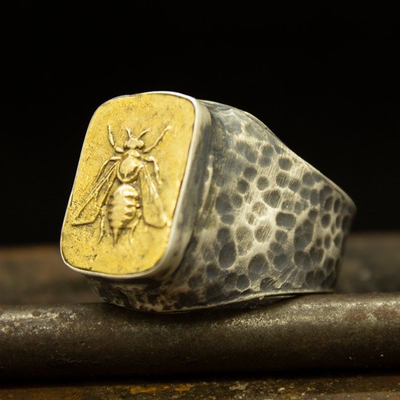 May include: A silver ring with a textured band and a square-shaped bezel featuring a gold bee design.