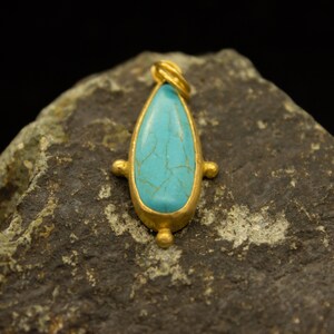 May include: A turquoise gemstone pendant with a gold setting and a teardrop shape. The pendant is sitting on a rough, gray rock.
