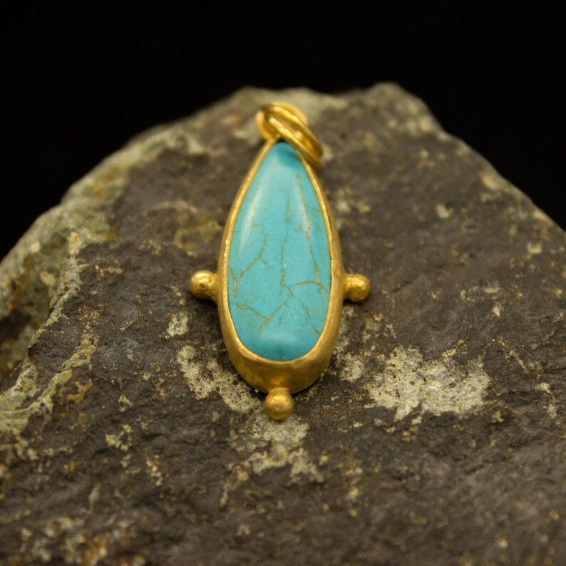 May include: A turquoise gemstone pendant with a gold setting and a teardrop shape. The pendant is sitting on a rough, gray rock.