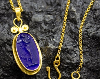 May include: A gold-colored necklace with an oval pendant featuring a deep blue cameo of a figure. The pendant is framed in gold and hangs from a delicate gold chain. The clasp is a simple hook and loop design.