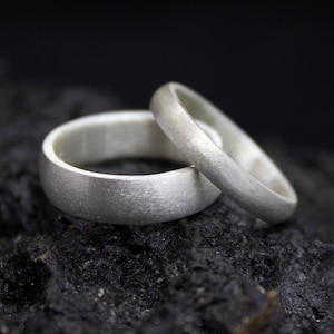 Può includere: Due fedi nuziali d'argento con finitura spazzolata, impilate l'una sull'altra. Le fedi sono su una superficie scura e testurizzata.