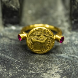 Puede incluir: Un anillo de oro con un dise&ntilde;o redondo, elevado, similar a una moneda que presenta un ciervo. El anillo tiene dos peque&ntilde;as piedras preciosas rosadas a cada lado de la moneda.