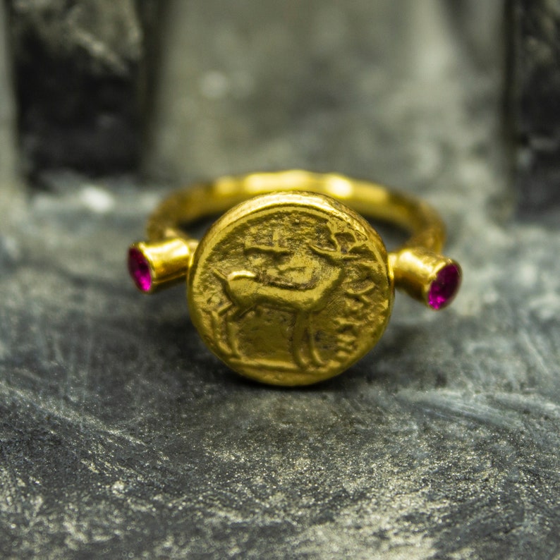 Puede incluir: Un anillo de oro con un dise&ntilde;o redondo, elevado, similar a una moneda que presenta un ciervo. El anillo tiene dos peque&ntilde;as piedras preciosas rosadas a cada lado de la moneda.