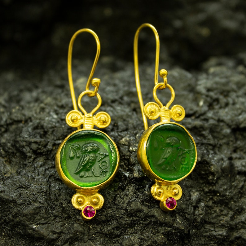 May include: Gold earrings with green glass owl charms and small red gemstones. The owl charms are detailed with a Greek design.